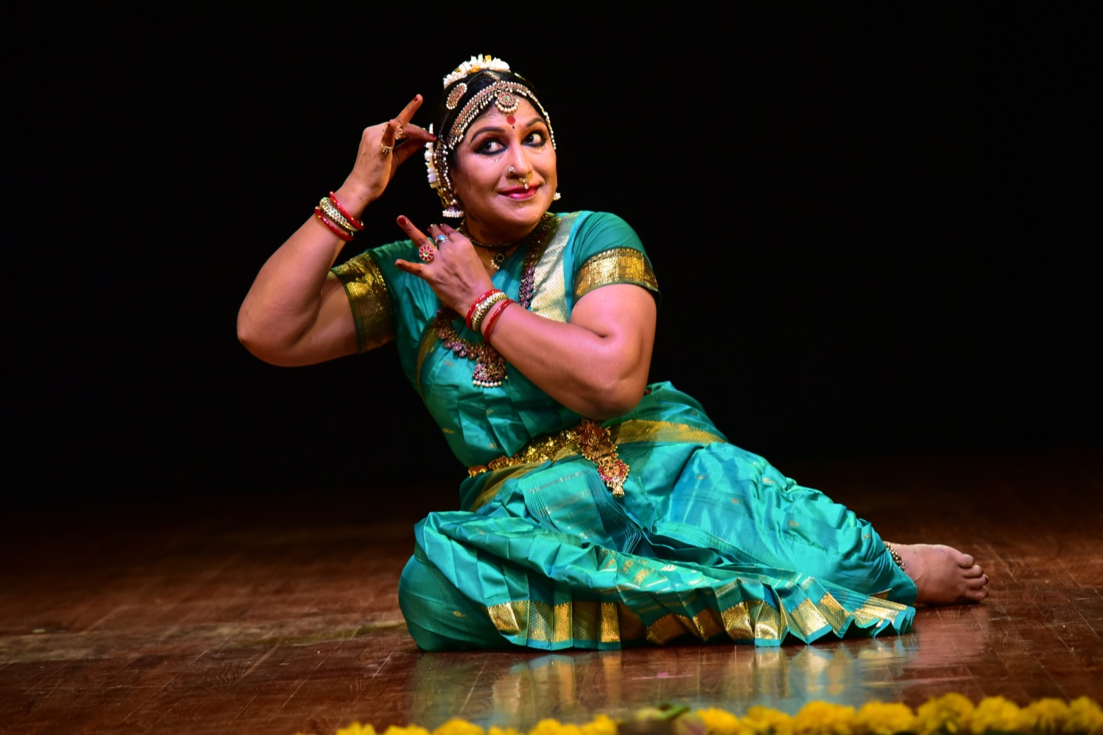 Narthaki Nataraj performing Bharatanatyam in a turquoise and gold costume, seated with expressive hand gestures
